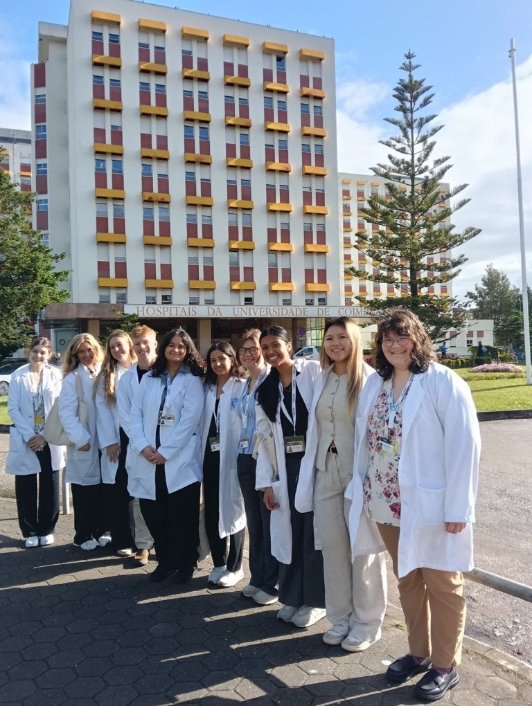 Group of students in white lab coats stand outside Coimbra University Hospitals building.