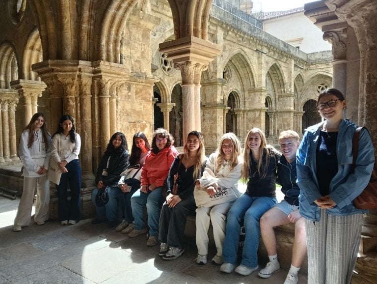 Group of students pose in a stone cloister with arched columns and vaulted walkways.