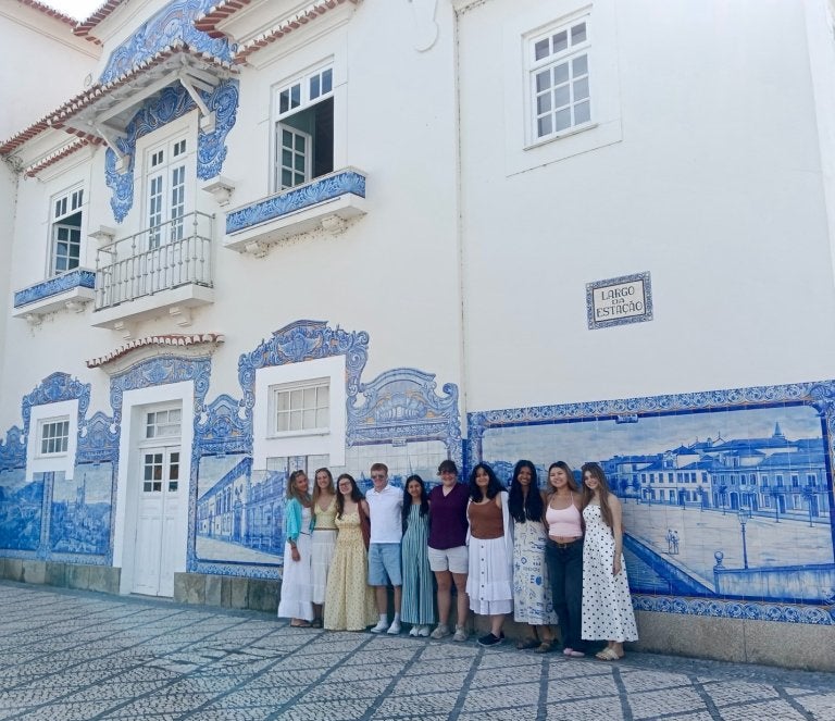 Group of students stand in front of a white building decorated with blue tile murals and balconies.