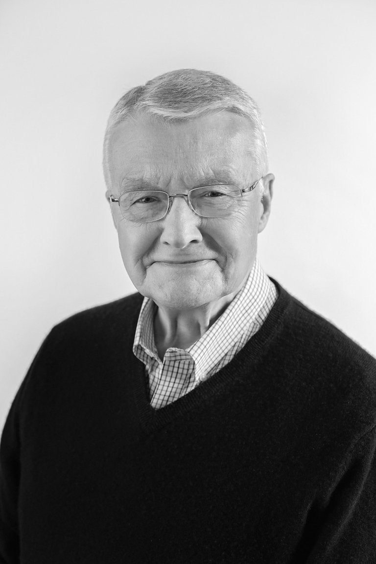 Person with short gray hair and glasses wears a dark sweater over a checkered shirt against a plain background.