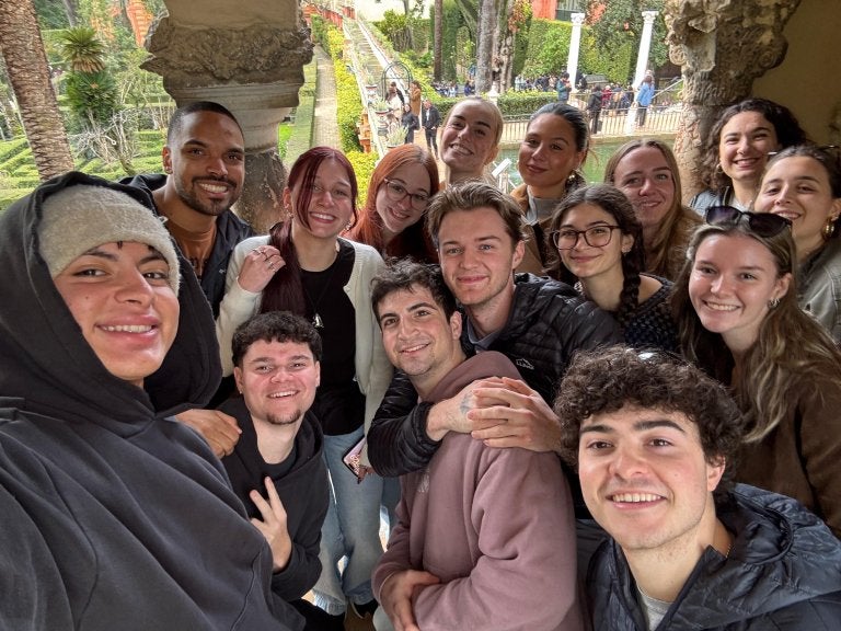 A group of people smile for a selfie under a stone archway with a formal garden in the background.