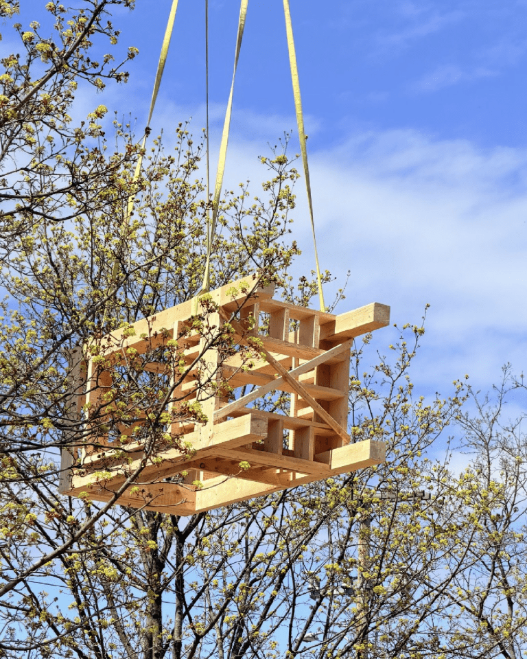 Wood framing for a small structure hangs from lifting straps above trees with spring blossoms.