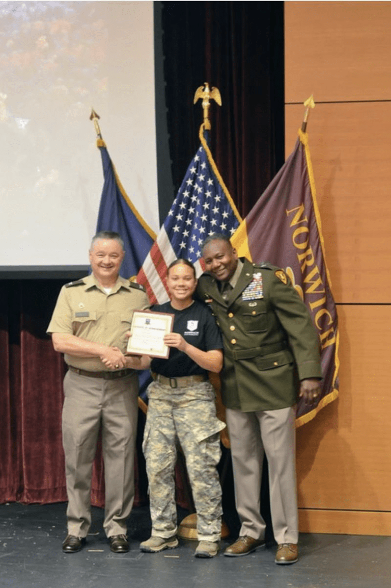 Cadet holds a certificate while standing with two uniformed service members in front of U.S. and Norwich flags.