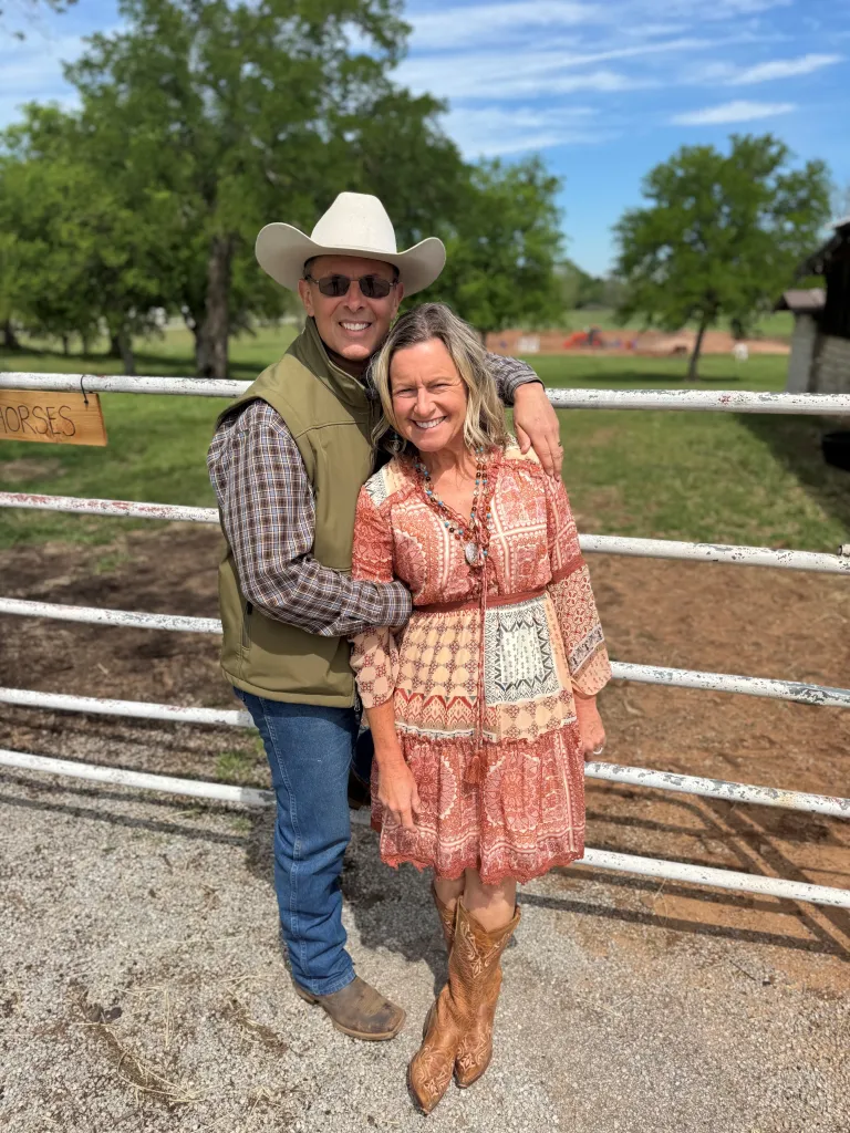 Two people stand together in front of a ranch gate, with one wearing a cowboy hat and both smiling at the camera.