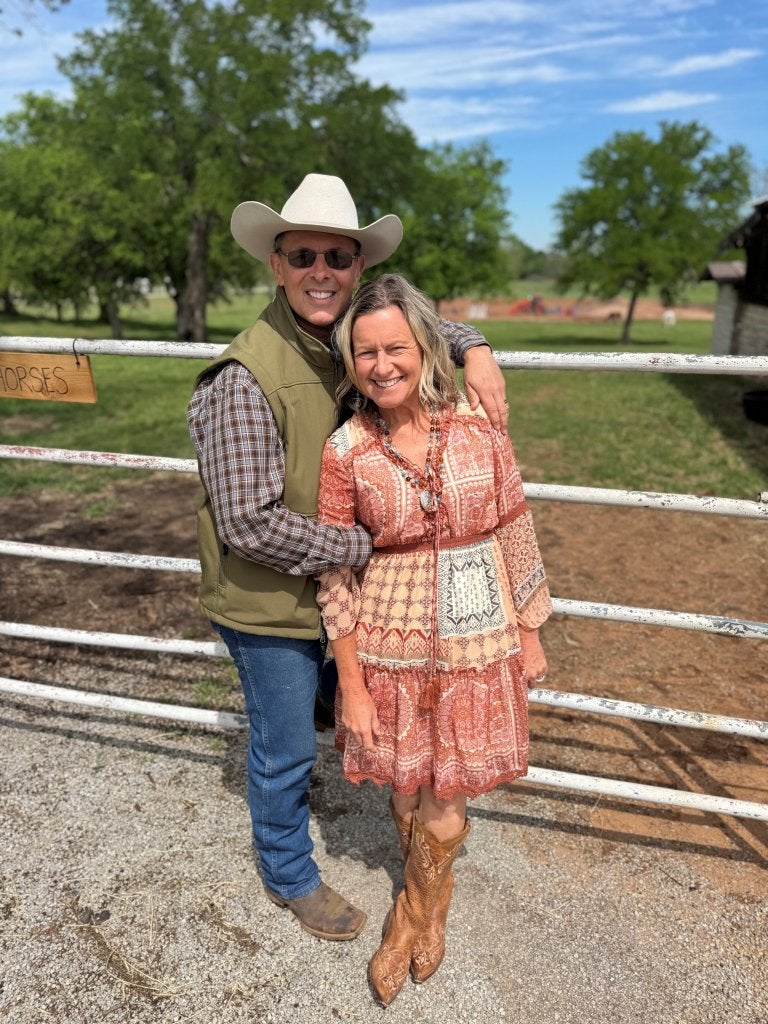 Two people stand together in front of a ranch gate, with one wearing a cowboy hat and both smiling at the camera.