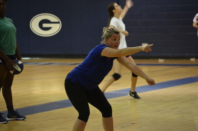 Volleyball coach points across the court while players practice in a gym.