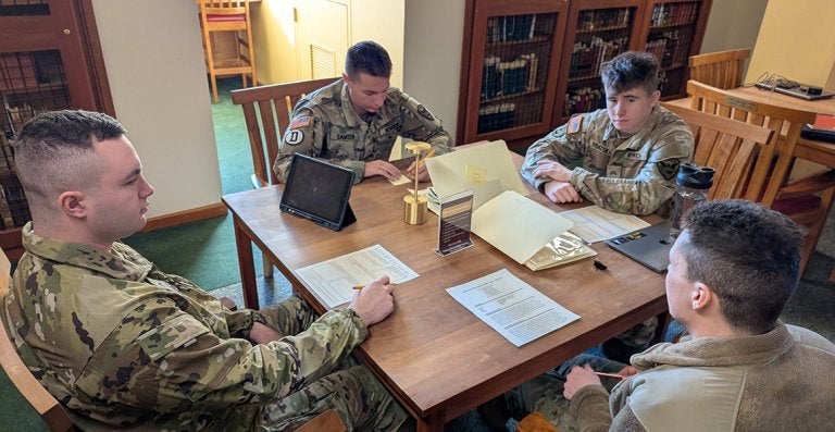 Four cadets in military uniforms study together at a library table with laptops, papers, and reference books.