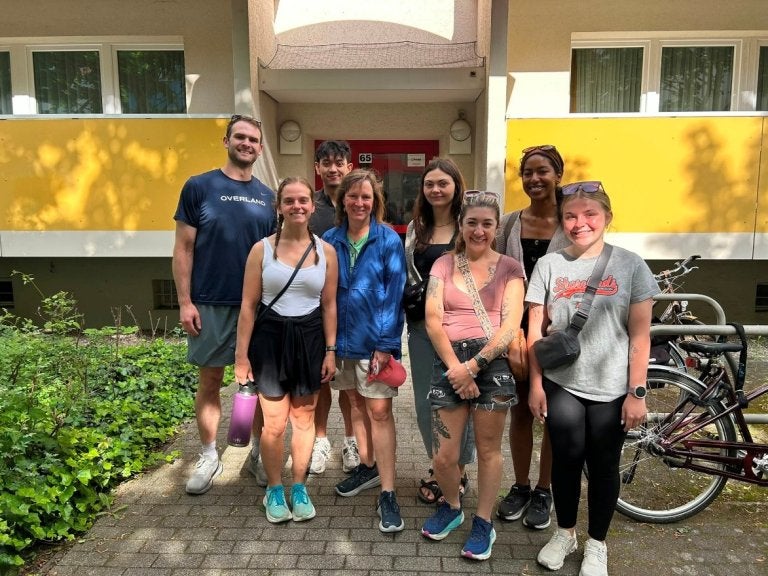 Eight people stand together on a walkway in front of an apartment building entrance with bicycles parked nearby.