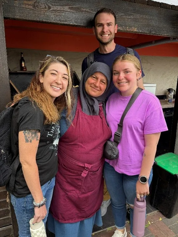 Four people stand together outside a food stand, with one person wearing a maroon apron and headscarf.