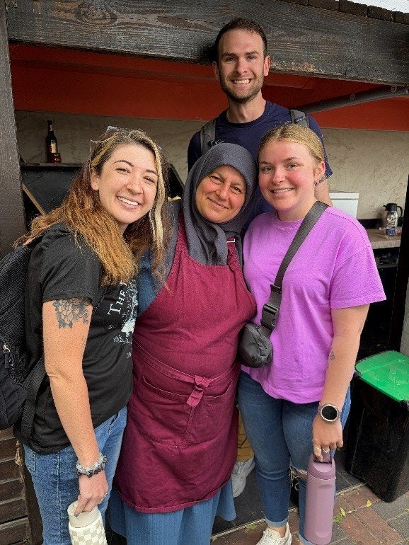 Four people stand together outside a food stand, with one person wearing a maroon apron and headscarf.