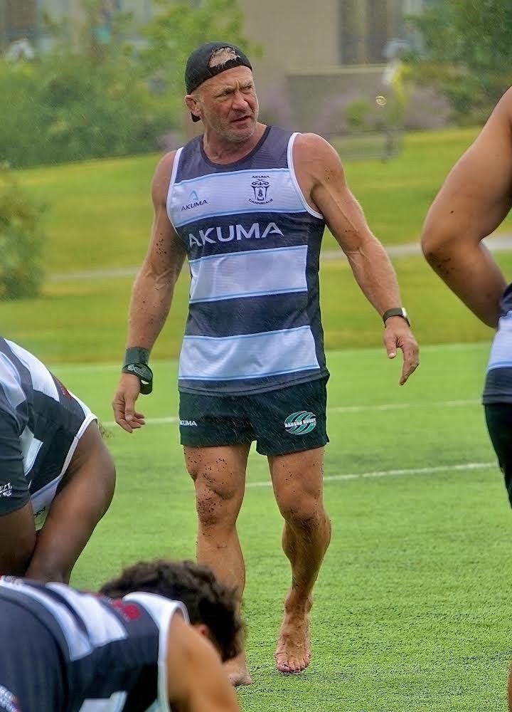 Rugby coach walks barefoot across a practice field while players stretch in the foreground.