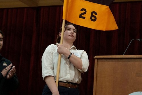 Person holds a black and yellow flag displaying the number 26 beside a podium on a stage.