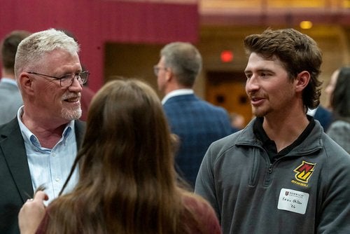 Two people wearing name tags talk at an indoor event while others stand and converse in the background.