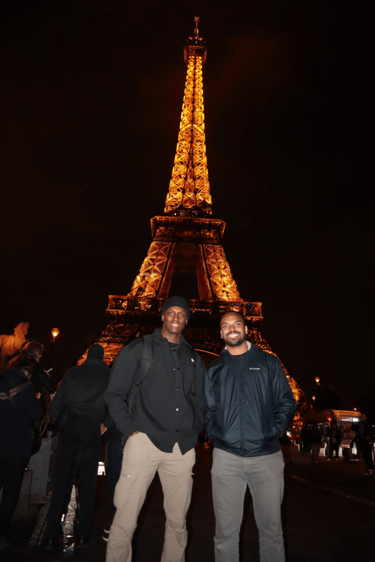 Two people stand in front of the illuminated Eiffel Tower at night.