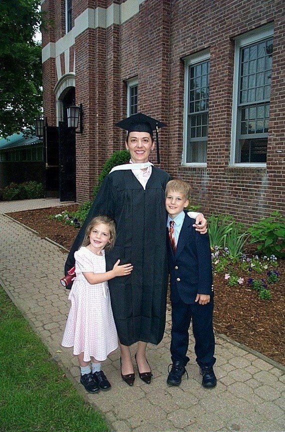Graduate in cap and gown stands with two children on a brick walkway outside a brick building.