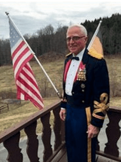 A man stands on a deck/porch with a US flag behind him.