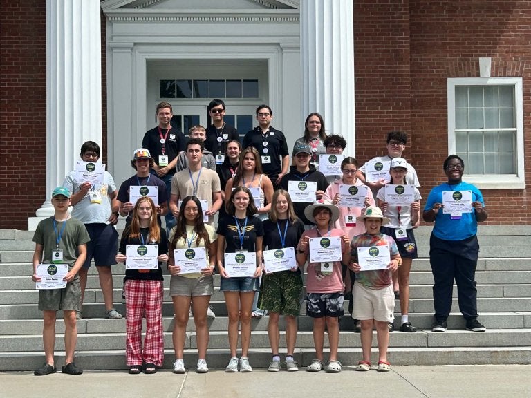 Group of students stand on building steps holding certificates in front of a brick building with white columns.
