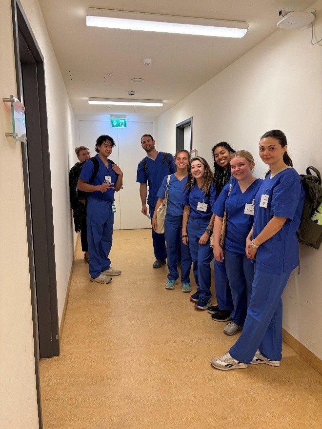 Seven people wearing blue medical scrubs stand in a hallway with ID badges visible.