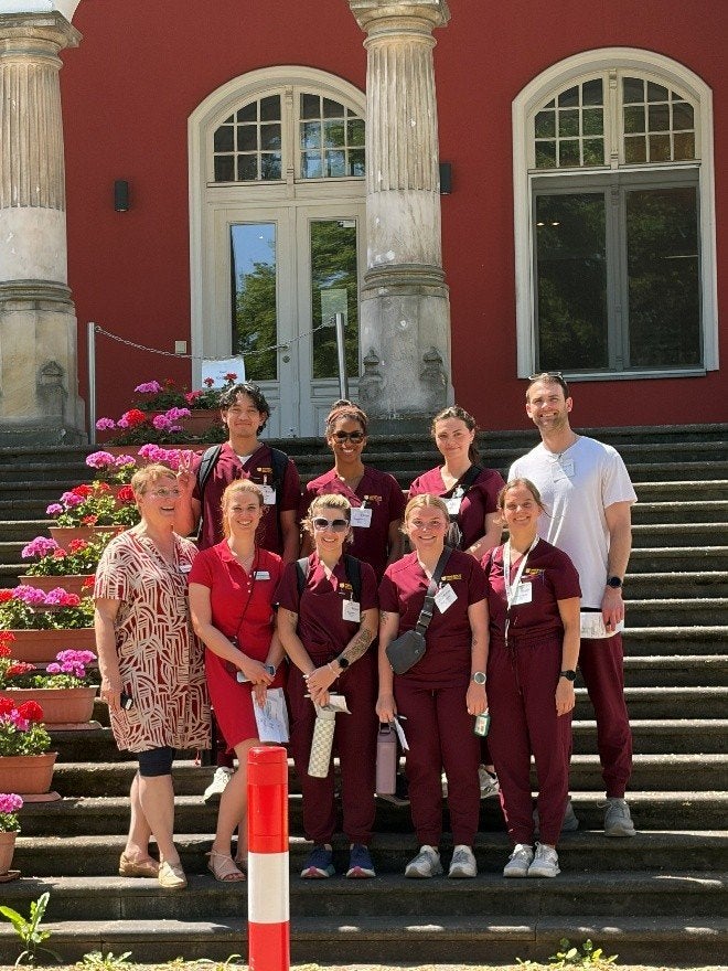 Nine people wearing medical scrubs and name badges stand on outdoor steps in front of a building with columns and flower pots.