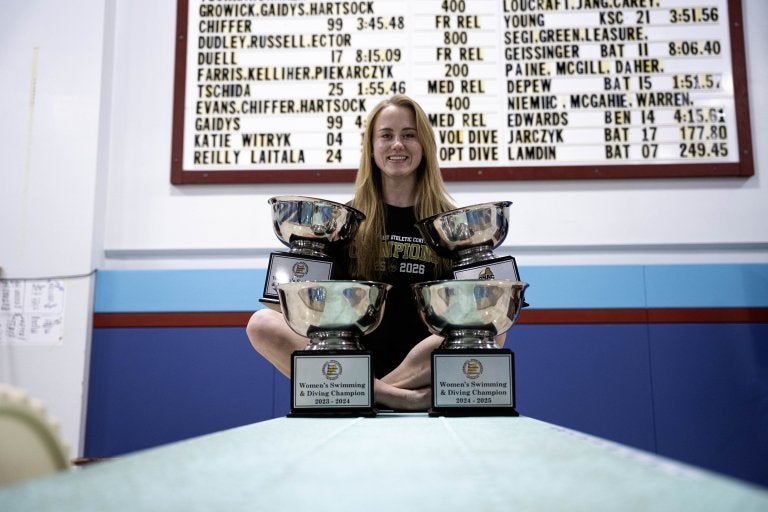 Swimmer sits behind four championship trophies beneath a scoreboard with event results.