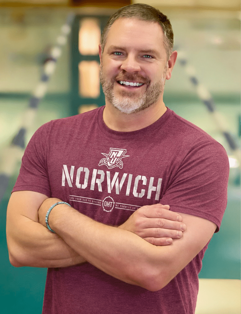 Coach stands with arms crossed wearing a Norwich T-shirt in front of an indoor pool.