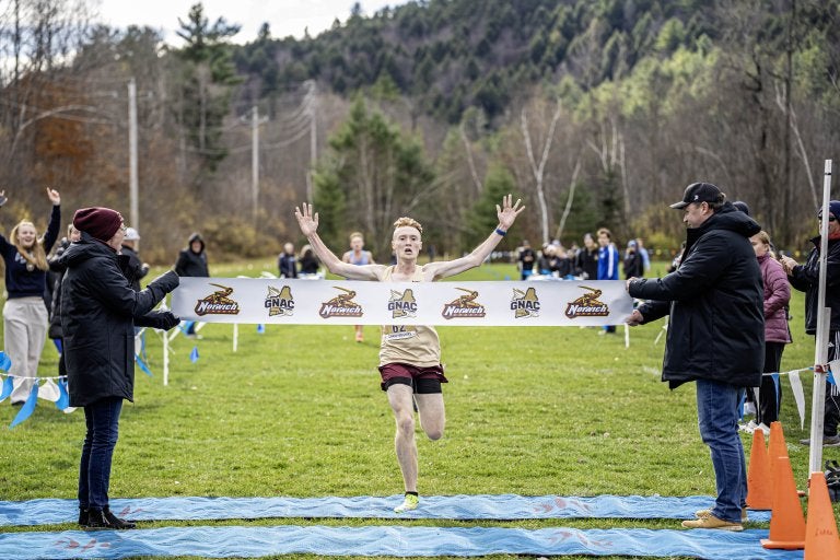 Cross country player breaks the finish tape with both arms raised as spectators line the course.