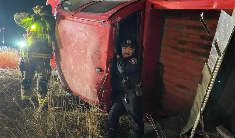 Police officer stands in the doorway of an overturned red vehicle while a firefighter works beside it at night.