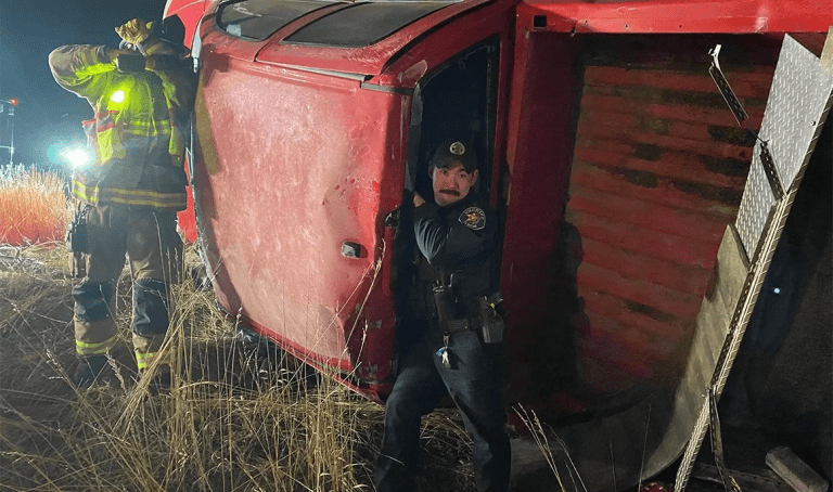 Police officer stands in the doorway of an overturned red vehicle while a firefighter works beside it at night.