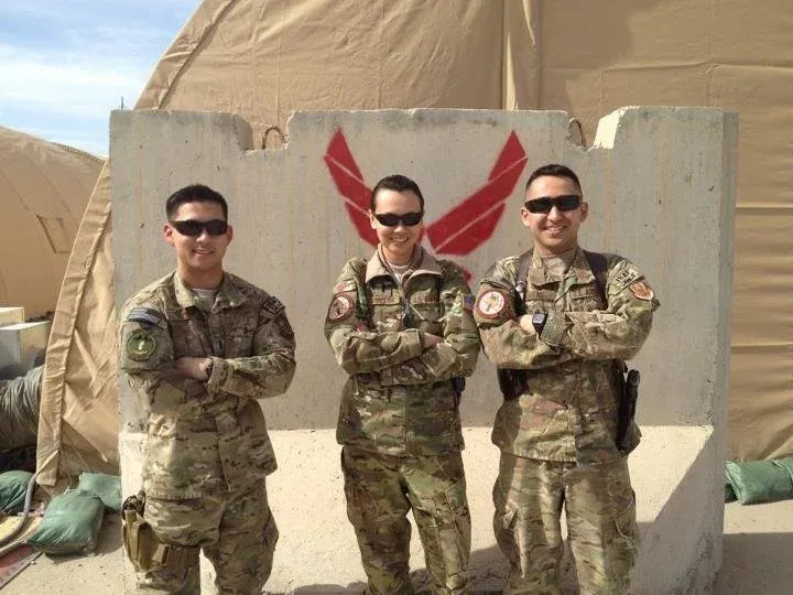 Three service members in camouflage uniforms stand with folded arms in front of a concrete barrier marked with a red Air Force symbol.