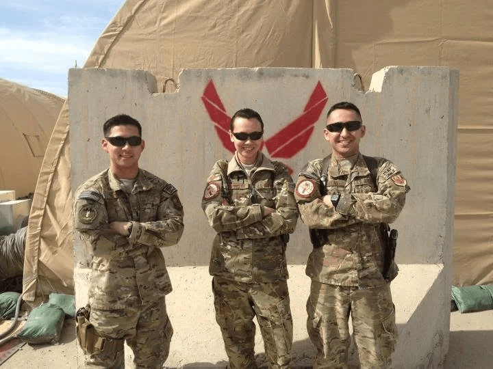 Three service members in camouflage uniforms stand with folded arms in front of a concrete barrier marked with a red Air Force symbol.