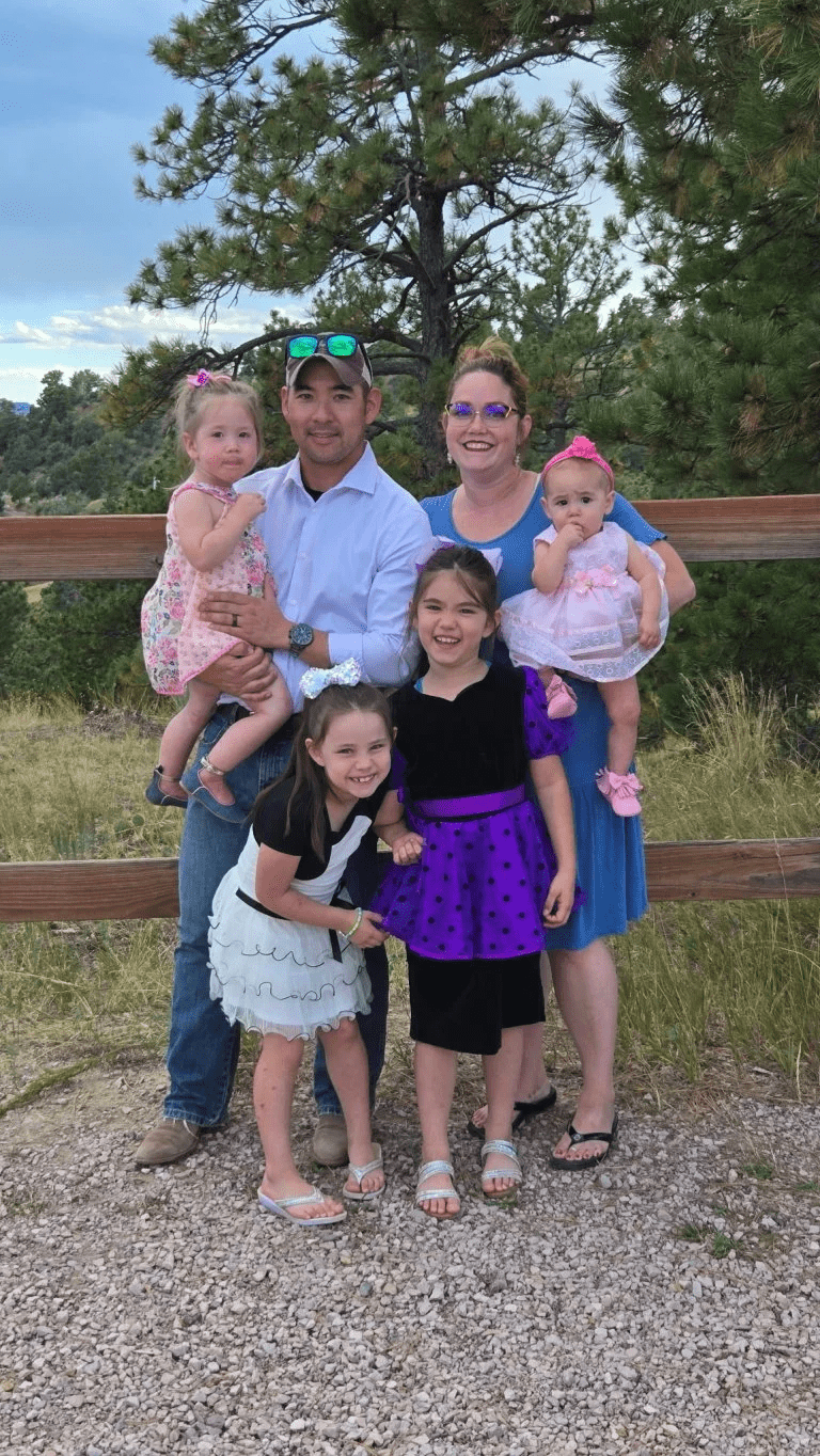 Two adults and four children pose together outdoors in front of a wooden fence and pine trees.