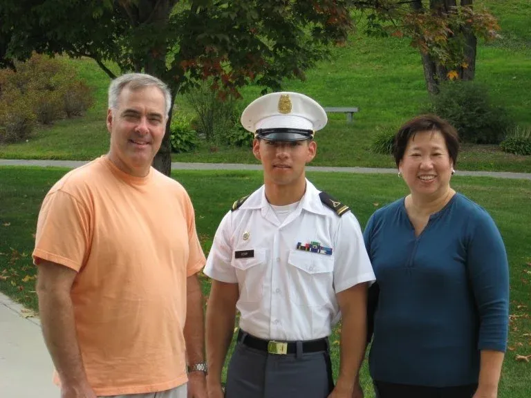 Cadet in white dress uniform stands between two adults on a campus lawn.