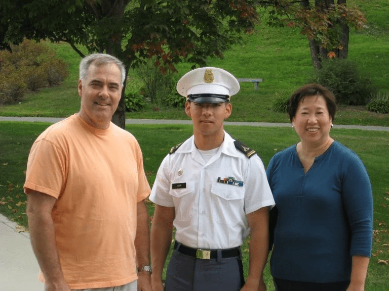 Cadet in white dress uniform stands between two adults on a campus lawn.
