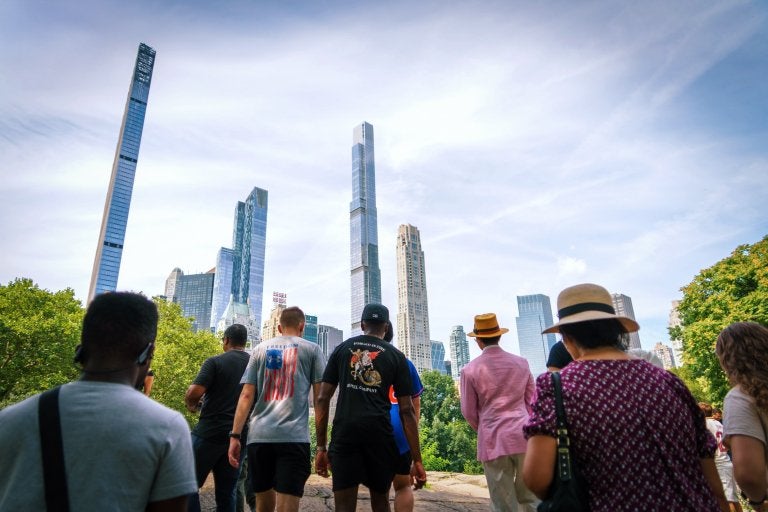 People walk through a park with tall city buildings in the background.