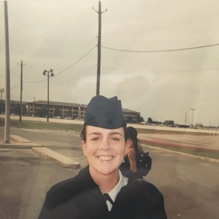 Service member in uniform and cap stands outdoors near a road and athletic field.