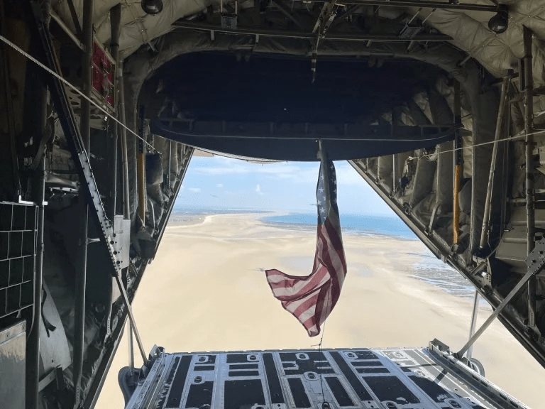 American flag hangs from the open rear of a military aircraft flying over a sandy coastline.