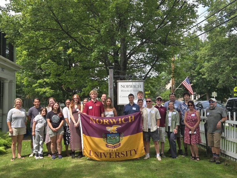 A group poses outdoors holding a Norwich University flag beside a Norwich Historical Society sign.