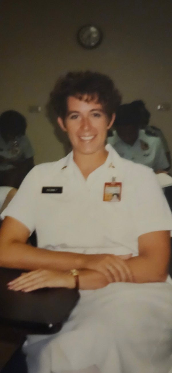 A service member in a white uniform sits with arms crossed indoors.