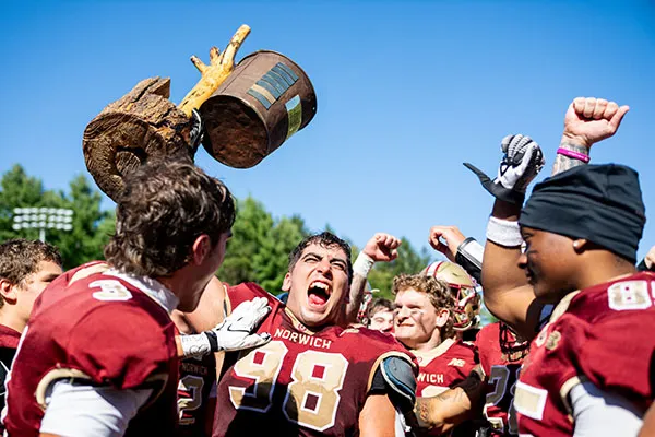 Norwich football players celebrate as one player holds a broken wooden trophy overhead.