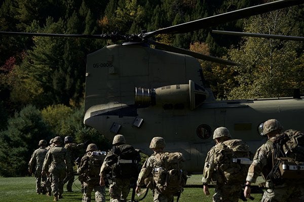 Service members carrying packs walk toward a military helicopter on a grassy field.