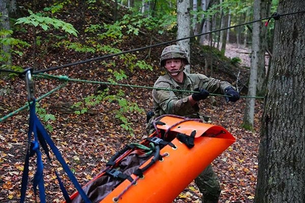 A service member in camouflage pulls a rope attached to an orange rescue litter in a wooded area.