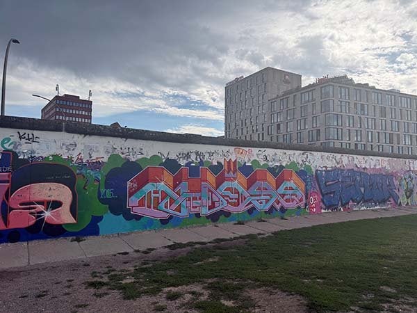 Long concrete wall covered in colorful graffiti with modern buildings and a cloudy sky behind it.