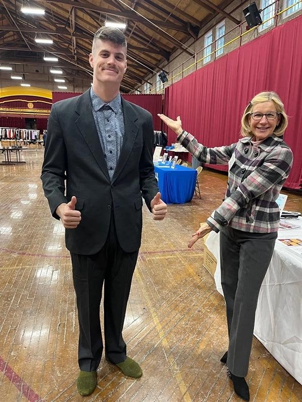 Person in a suit gives two thumbs up as another person gestures toward them in a large hall.