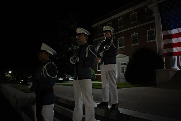Three cadets in uniform hold bugles on steps beside a large American flag at night.