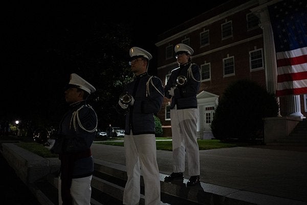 Three cadets in uniform hold bugles on steps beside a large American flag at night.