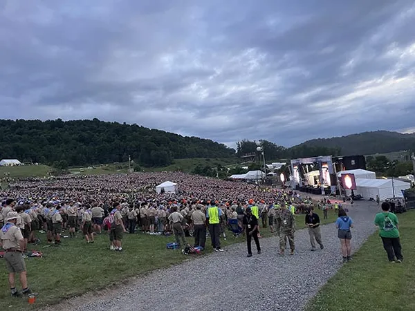 Large crowd gathers on a grassy hillside facing an outdoor stage under a cloudy sky.