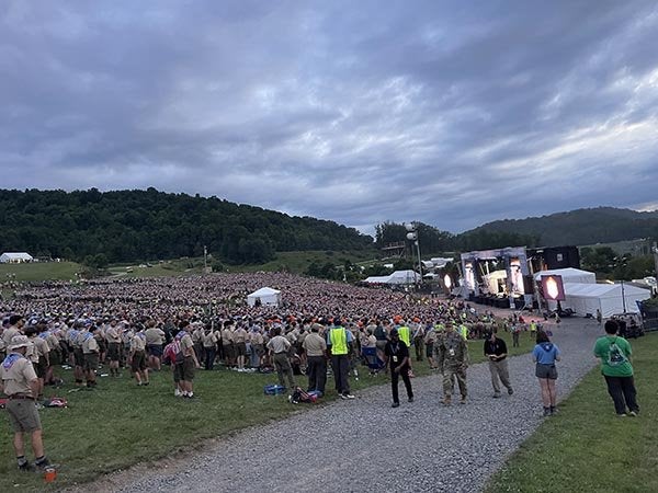 Large crowd gathers on a grassy hillside facing an outdoor stage under a cloudy sky.