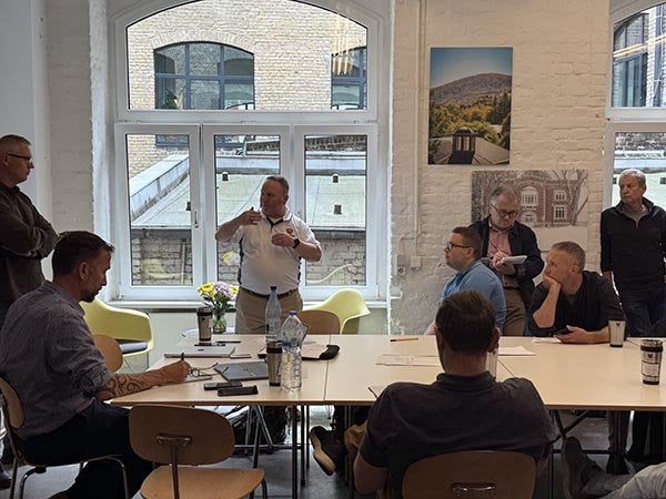 A person speaks to a group seated around tables in a bright room with large windows.