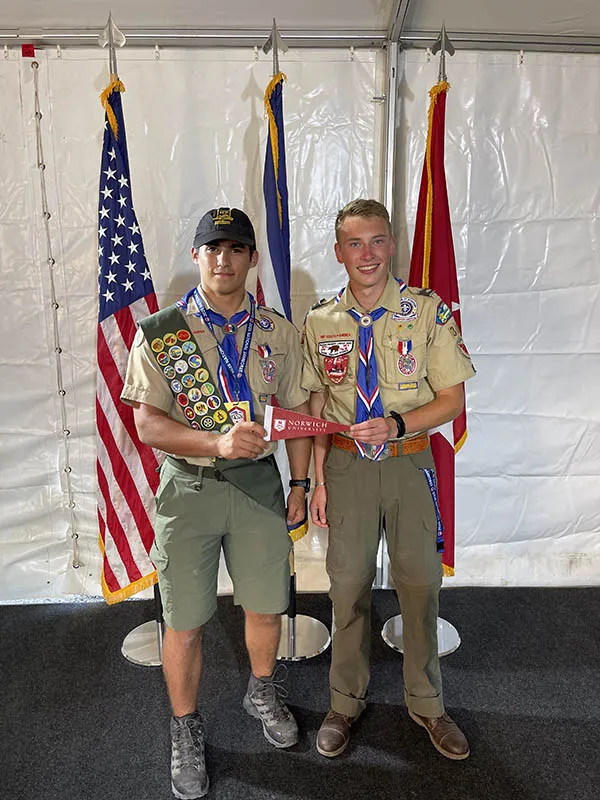 Two scouts in uniform hold a Norwich University pennant in front of three flags.