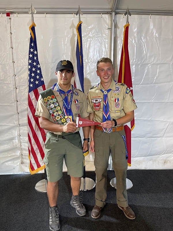 Two scouts in uniform hold a Norwich University pennant in front of three flags.